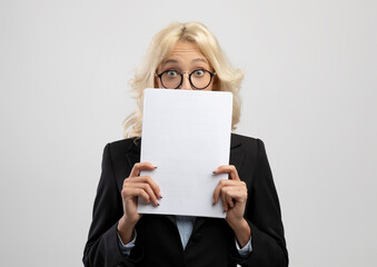 Shocked female entrepreneur peeking from behind documents, posing over light grey studio background
