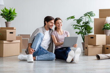 Glad millennial caucasian family in casual searching ideas for new interior on computer, sits among cardboard boxes - obrazy, fototapety, plakaty