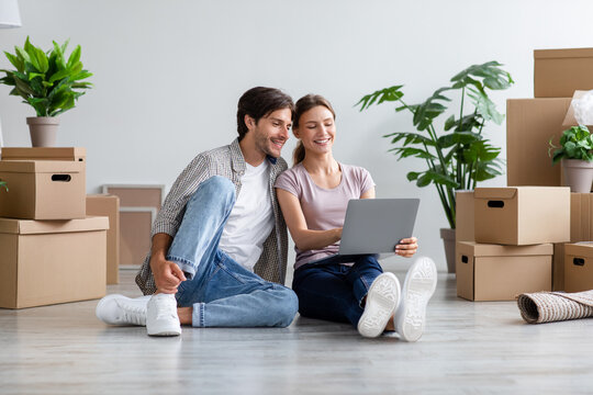 Glad millennial caucasian family in casual searching ideas for new interior on computer, sits among cardboard boxes