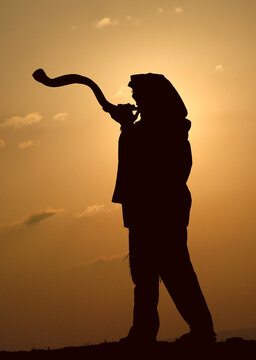 Silhouette of the entire body of a Jewish man wearing a tallit and tefillin and blowing a long shofar made from the horn of a kudu antelope at sunrise during morning prayers  in Israel.