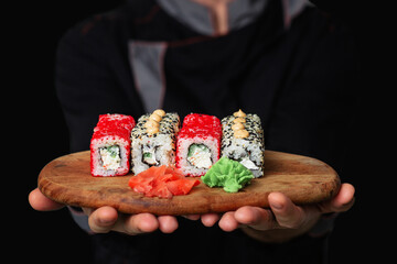 Close-up of the hands of a Man chef in a black uniform holding a wooden tray plate full of sushi wasabi ginger