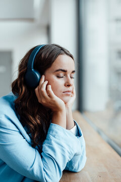 Young businesswoman with head in hands listening music through wireless headphones at desk