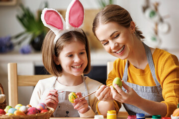 Loving young mother teaching happy   kid daughter to decorate Easter eggs while sitting in kitchen