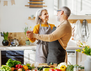 Happy senior elderly couple husband and wife embracing and dancing while cooking together in kitchen
