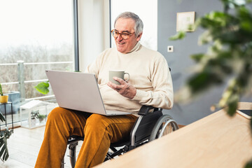 Smiling disabled senior man holding coffee cup using laptop sitting on wheelchair at home