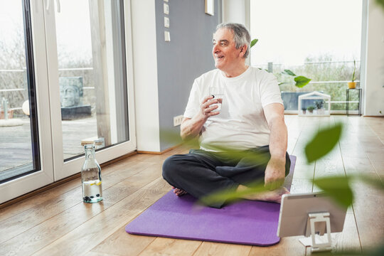 Smiling senior man with drinking glass sitting cross-legged on exercise mat at home