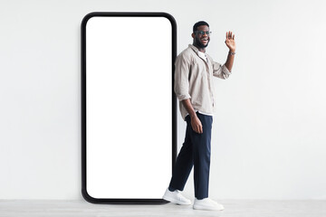 Happy black guy standing next to giant smartphone with white screen and waving hello to camera,...