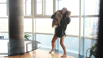 Beautiful dark-haired girl in pajamas dancing with a Maine Coon cat at home in the living room.