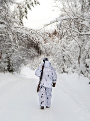 A male hunter in camouflage, armed with a rifle, stands with his back in a snowy winter forest.