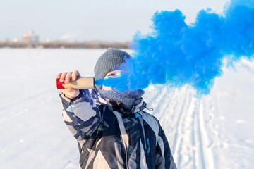 A male teenager in winter in an open space holds a smoke bomb with blue smoke in his hands. The concept of freedom and rebellion.
