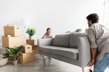 Smiling millennial caucasian couple in casual carry sofa in room with packed cardboard boxes, planning interior