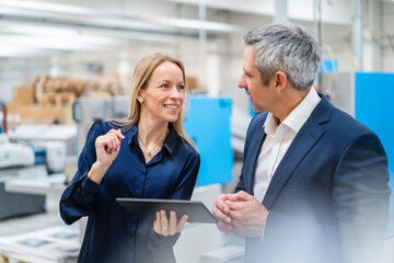 Happy blond businesswoman holding tablet PC discussing with colleague in factory