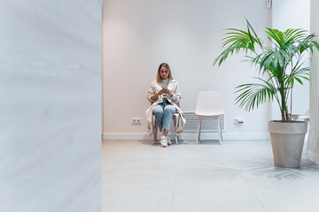 Young woman using mobile phone sitting in waiting room at clinic