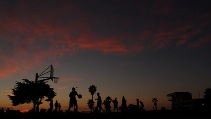 Fototapeta na wymiar Silhouettes of players on basketball court outdoor, people playing basket ball game, sunset ocean beach, California coast, USA. Black hoop, net and backboard on streetball sport field. Mission beach.