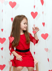 A girl with loose long hair in a red dress sits on a stool against a white background with hearts. Concept Valentines Day.