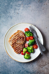 Grilled pork steak with broccoli and tomatoes on plate, dark background.