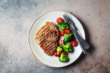 Grilled pork steak with broccoli and tomatoes on plate, dark background.