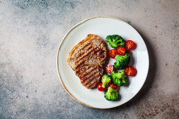 Grilled pork steak with broccoli and tomatoes on plate, dark background.