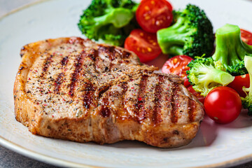 Grilled pork steak with broccoli and tomatoes on plate, dark background.