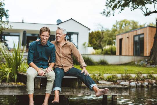 Happy father and son sitting on jetty by lake at backyard - Powered by Adobe