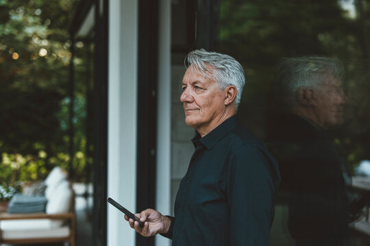 Senior man with mobile phone standing by glass wall