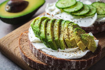 Toasts with cream cheese, avocado, cucumber and rye bread on wooden board, dark background.