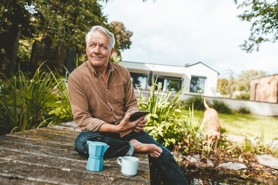 Smiling senior man with tablet PC sitting on jetty at backyard - Powered by Adobe