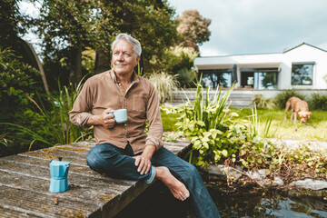 Smiling senior man with coffee cup sitting on jetty by lake at backyard