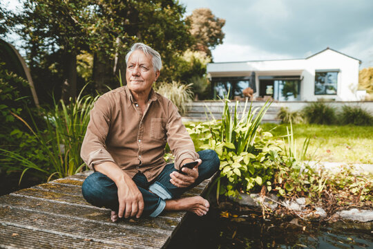 Senior man with smart phone sitting cross-legged on jetty by lake