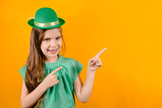 Funny girl child in a hat of a leprechaun for a St. Patrick's Day isolated on a yellow background. Traditions, technologies, holidays concept. Kid points with his fingers to the place for the text.