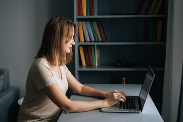 Side view of focused business woman working on laptop computer from home office sitting at desk in dark room. Serious businesswoman studying online, web surfing information, shopping in internet store