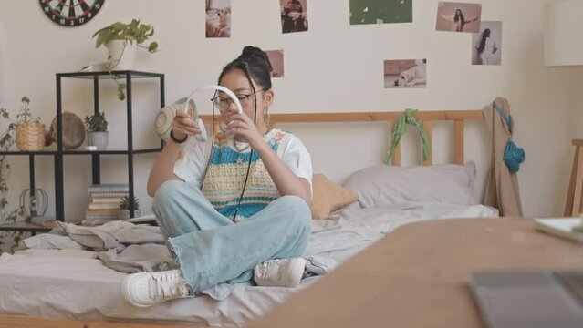 Tracking shot of Asian teenage girl in hipster outfit taking smartphone from table, putting on headphones and starting listening to music sitting on bed in her room
