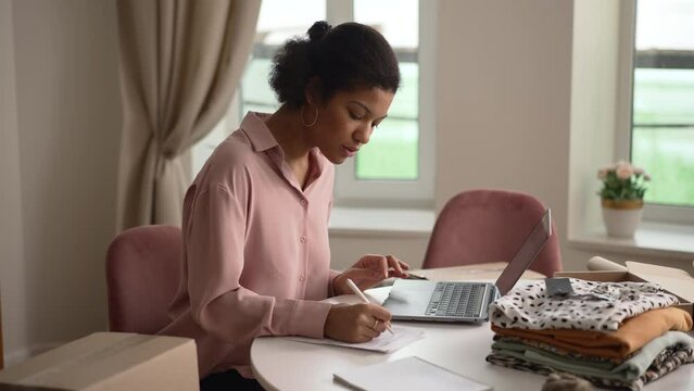 Young woman writing return order and looking at laptop while sitting at table at home spbd. 4k video Beautiful african american female consumer browses web on computer and writes on paper, wants to