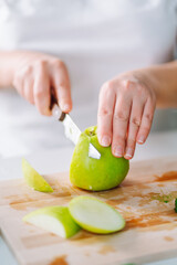 Cropped shot of woman in white t-shirt cutting fresh green apple on wooden chopping board, preparing healthy diet food, cooking vegan fruit and vegetable salad behind table in kitchen, selective focus