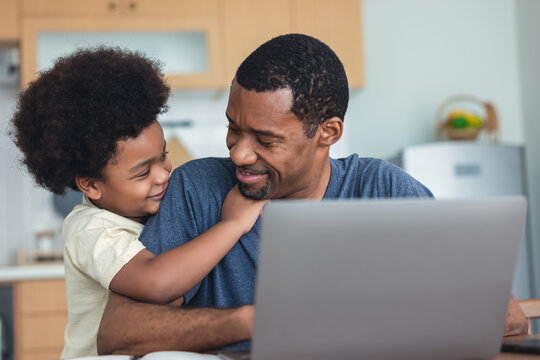 Portrait of African american cute little boy hugging of handsome dad working laptop computer while sitting at kitchen table at home. African-american family spending time at home with kid.