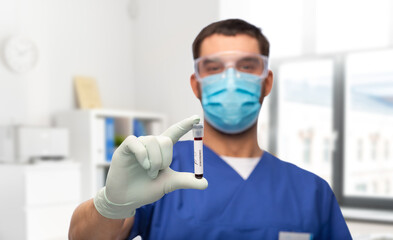 healthcare, coronavirus and medicine concept - doctor in blue uniform, face protective medical mask, goggles and gloves with blood in test tube over medical office at hospital background