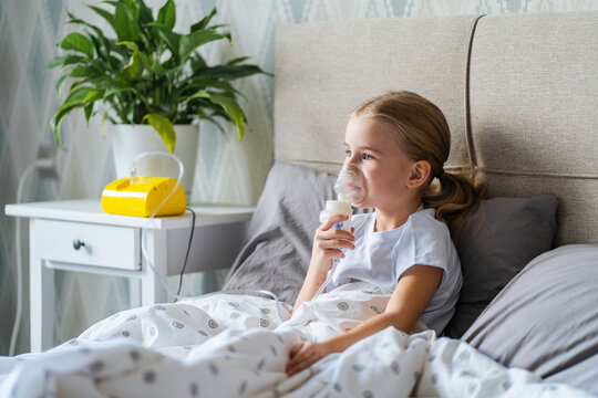 Sick little girl making inhalation with nebulizer to reduce coughing, lying in bed at home, child taking medication while breathing in through face mask. Bronchitis and asthma treatments for children