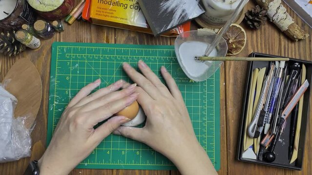 Woman makes egg mold out of white clay. Tools for modeling and design of ceramic souvenirs. Hobbies, handmade and crafts. Water, stationery knife, sticks on wooden table.