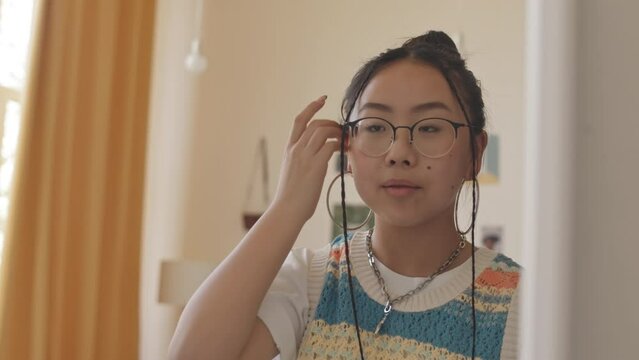 Chest up shot of pretty Asian teenage girl in eyeglasses looking at herself in mirror
