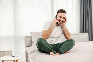 Fototapeta premium A happy morning man talking on the phone while sitting on the bed in the morning at his cozy home.