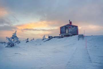 Cabin in winter dawn. Snow-covered staircase leading through snowdrifts to a lonely house on a hilltop in the cool evening.