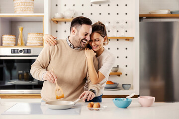 A man in the kitchen preparing a meal while his wife hugging him.