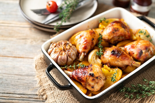 Baked chicken legs and potatoes with oranges, garlic and onions in a baking dish on a light gray wooden culinary background closeup	