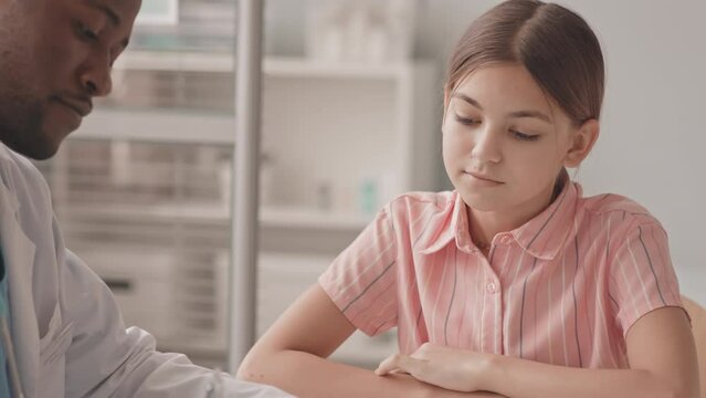 Waist-up slowmo shot of young African-American pediatrician or ent-doctor examining lymph nodes of 13-year-old Caucasian schoolgirl having medical check-up in clinic