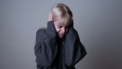 Isolated Cute Young Caucasian Girl Standing on a Grey Coloured Background, Covering Her Ears, Looking Like She is in a Lot of Pain. Wearing a Comfy Black Sweater. Depressed Female, Mental Health.
