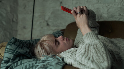 Depressed Young Blonde Woman Texting on Her Phone, Refusing to Get Up From the Sofa. Beautiful Girl Looking Sad While Laying on the Couch on Her Smartphone.
