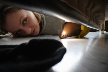Woman cleaner looking under sofa for sock using flashlight - obrazy, fototapety, plakaty