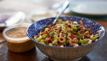 A bowl of traditional healthy Greek salad with olives, feta cheese, cucumber and peppers with a hummus dip beside it.