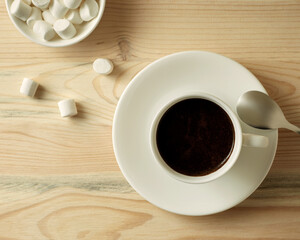A cup of black coffee on a wooden table. Next to the cup is a small plate of marshmallows. View from above
