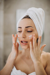 Young woman applying face cream on her face with towel on head